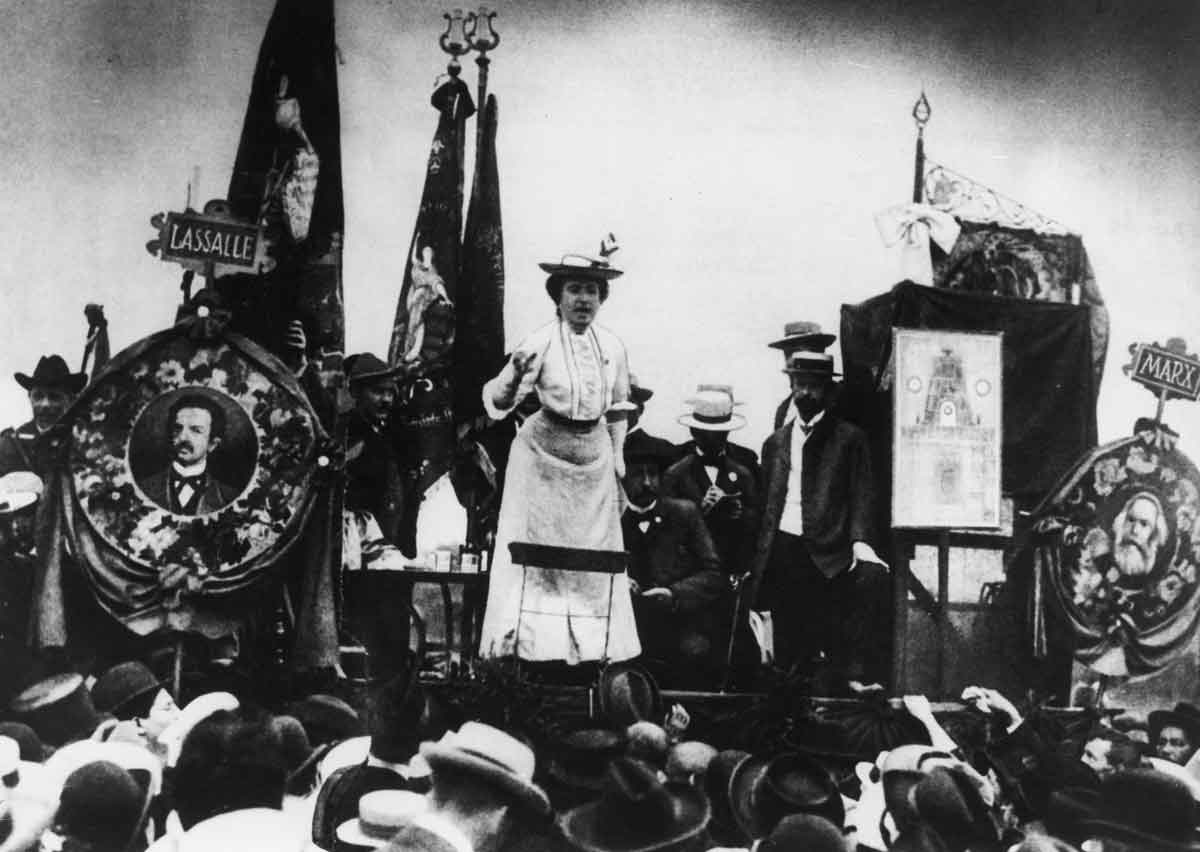A black and white image of Rosa Luxemburg speaking to a crowd from a podium.