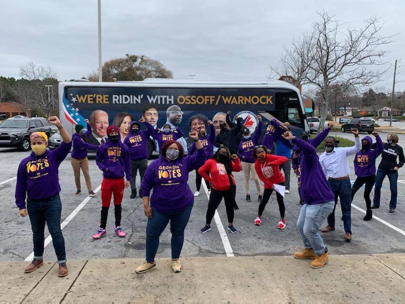 A group of volunteers in matching t-shirts stand in front of a campaign bus for the Working Fam