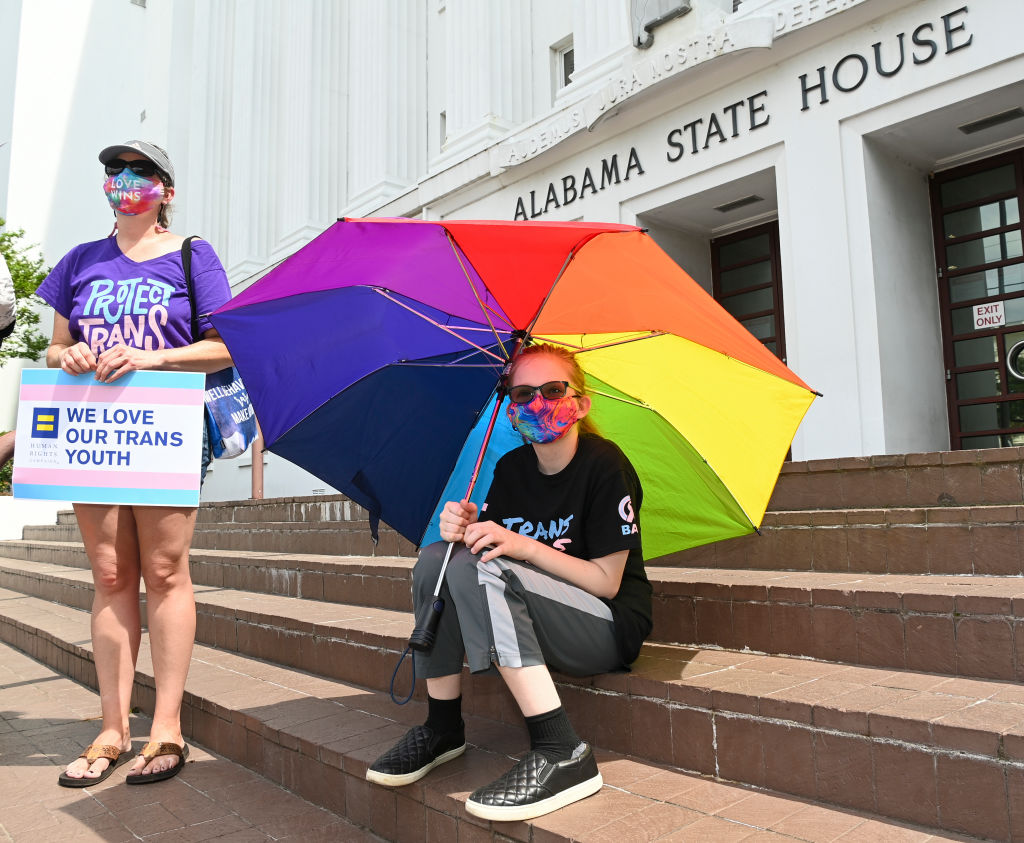 Two women are in front of the Alabama State House, one seating on the stairs and holding a rainbow umbrella, the other standing holding a sign that says "