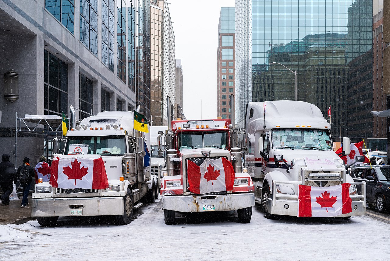 Three large trucks on a snowy city street with Canadian flags attached to their fronts.
