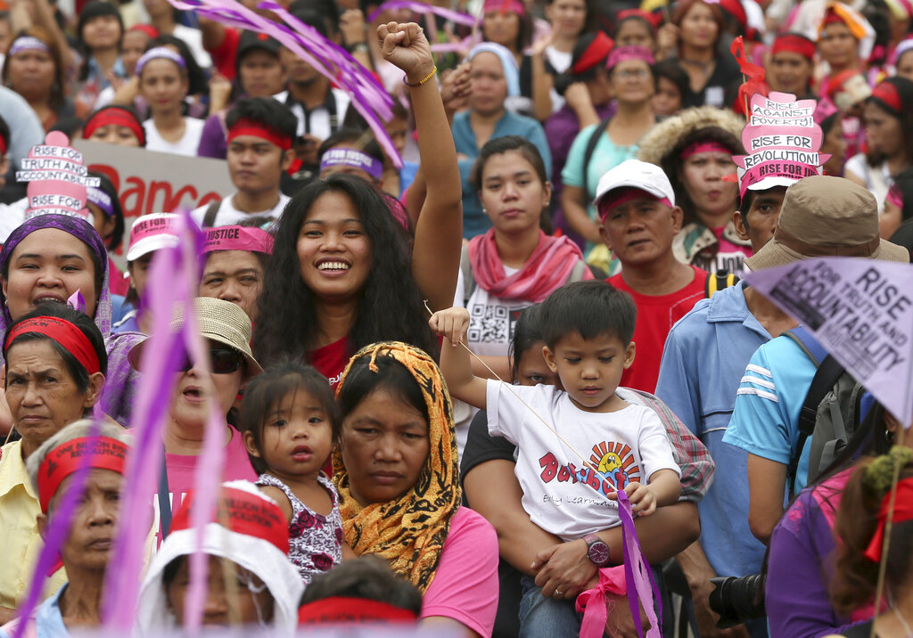 A large group of people, mostly women and children, is facing the camera during a demonstration in Manila. Some are wearing red headbands and purple flags. A woman with long black hair is a bit higher than the rest and has her right fist up. In front of her, a woman with hijab holds a toddler in her arms.